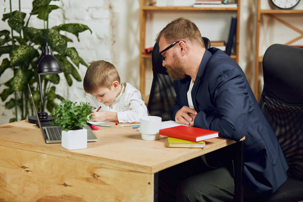 Mature man, father sitting at table, working remotely at home and doing homework with his little son. Attentive parent. Concept of fatherhood, childhood, family, freelance job, home office - Photo, image