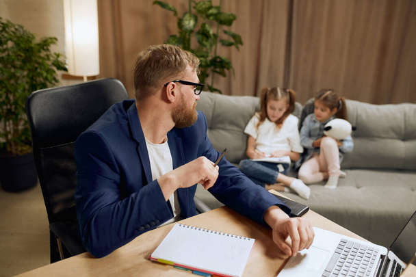 Dospělý muž, otec sedící u stolu a pracující na notebooku doma a starající se o své malé děti hrající si na gauči. Pojem otcovství, dětství, rodina, práce na volné noze, domácí kancelář - Fotografie, Obrázek