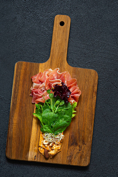 Salad of jamon (prosciutto) spinach and lettuce with crispy savory bread, nuts, microgreens on black background. Flat lay of food in form of light bulb on board. Food energy concept, top view. - Фото, зображення