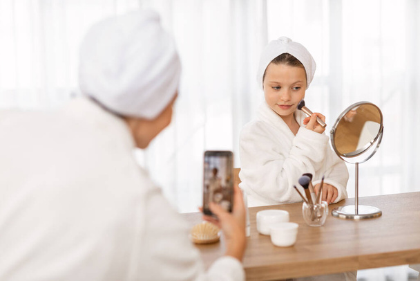 Beauty Tutorial. Mother Recording Video Of Her Daughter Doing Makeup At Home, Mom And Female Child Wearing Bathrobes And Towels On Head Capturing Content For Social Media, Selective Focus - Foto, Bild