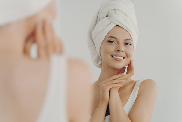 Kaunis nuori nainen suihkun jälkeen näyttää peilistä varovasti koskettaa pehmeä terve hehkuva kasvot iho, onnellinen tuhatvuotinen nainen soveltamalla kauneudenhoitotuotteita aikana päivittäin kasvojen menettelyt, ihonhoito käsite - Valokuva, kuva