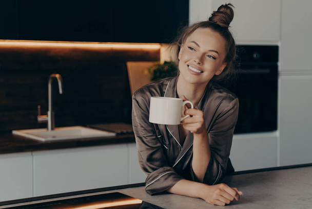 Onnellinen hymyilevä nuori nainen kotivaatteissa nauttimassa aromaattista aamukahvia modernissa keittiössä heräämisen jälkeen ennen kiireistä työpäivän alkua, positiivinen nainen viettää vapaa-aikaa kotona. - Valokuva, kuva