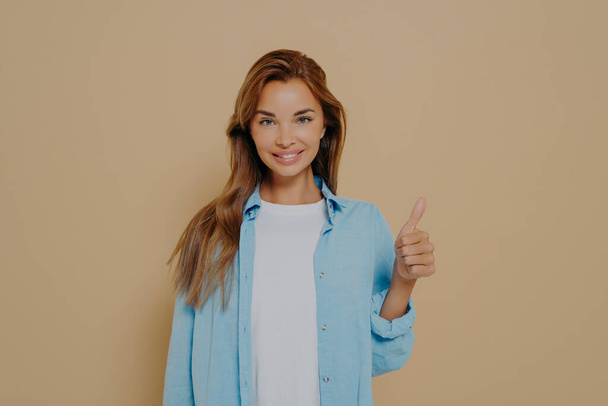 Happy young caucasian female in blue shirt with long sleeves rolled up weared over white tshirt making thumb up sign and smiling on camera with support on isolated pastel beige colored background - Foto, Bild