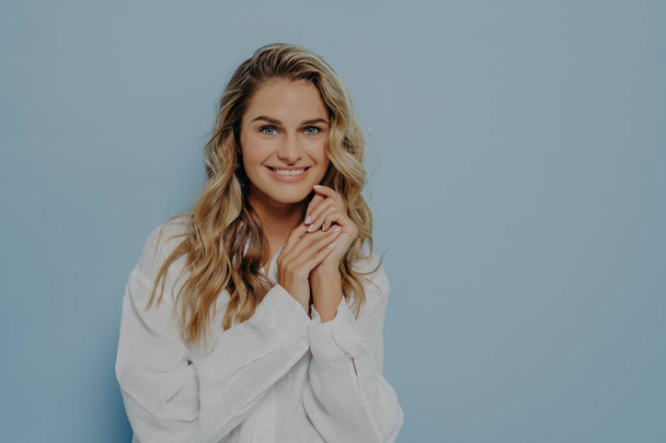 Pleased young caucasian woman with blonde wavy hair wearing white casual shirt smiling broadly and keeping hands near face, beautiful female glad to hear good positive news. Happy people concept - Photo, Image