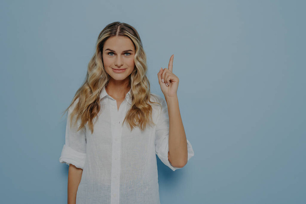 Calm blonde girl with slight smile pointing up in air with her finger, has new brilliant idea and showing it with gesture while standing next to blue wall. Female showing at copy space - Photo, Image