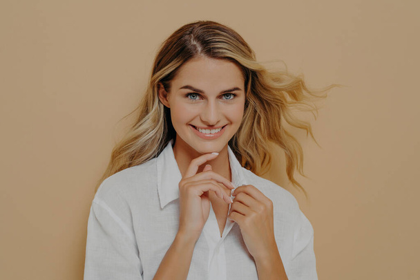 Close up portrait of flirty romantic and feminine beautiful blond woman looking at camera playfully and with bright wide smile, holding hands in front of her, isolated over beige studio background - Foto, imagen