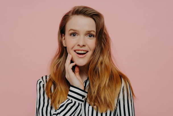 Amused young woman in striped black and white blouse with wavy ginger medium length hair touching face, expressing amazement while posing over light pink studio wall. Human emotions concept - Photo, image
