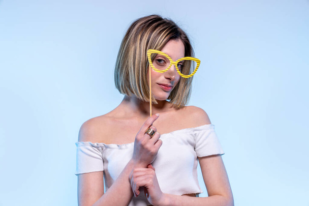 Young caucasian girl with light blue eyes looks in camera with funny glasses - Studio photography of pretty woman having fun with fake eyewear - 写真・画像