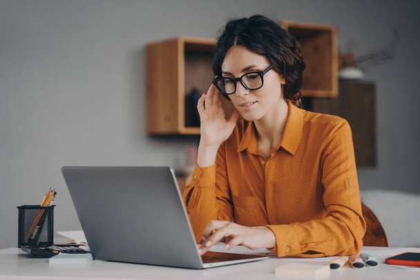 Freelance. Beautiful focused Spanish woman in glasses sits at desk in living room types communicates with client live on laptop, focused young female consultant adviser working remotely online at home - Photo, Image