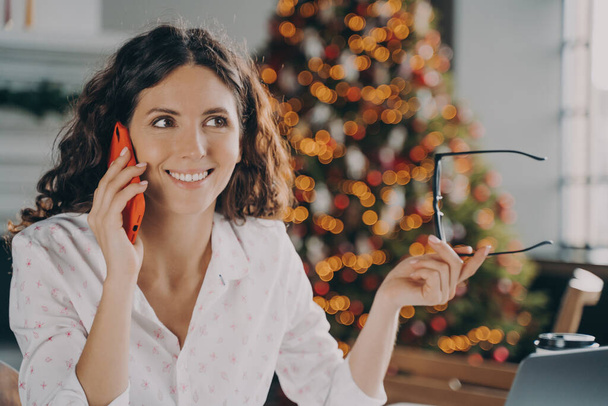 Νεαρή ευτυχισμένη Ευρωπαία επιχειρηματίας που μιλάει στο κινητό τηλέφωνο με πελάτη ενώ κάθεται στο χώρο εργασίας με διακοσμημένο χριστουγεννιάτικο δέντρο πίσω, χαμογελαστή γυναίκα που εργάζεται στο γραφείο στο σπίτι κατά τη διάρκεια των χειμερινών διακοπών - Φωτογραφία, εικόνα