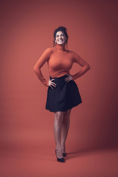 Ενθουσιασμένη κοπέλα χαμογελά διασκεδάζοντας με πορτοκαλί φόντο, πουλόβερ και μαύρη φούστα - Φωτογραφία, εικόνα