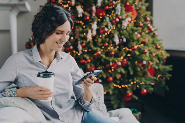 Usmívající se evropská žena si užívá novoroční dopoledne doma, šťastná hispánka s mobilním telefonem a šálkem kávy sedí u vánočního stromku, používá smartphone, píše zprávu s přáním dovolené - Fotografie, Obrázek