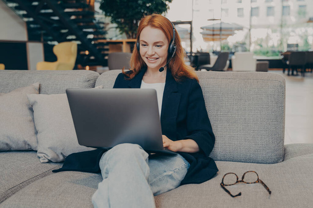 Uśmiechnięta biała kobieta freelancer z rudymi włosami nosząca bezprzewodowe słuchawki, komunikująca się z klientem przez telefon wideo na laptopie, dająca lekcję języka online, siedząca na wygodnej sofie w lokalnej kawiarni - Zdjęcie, obraz