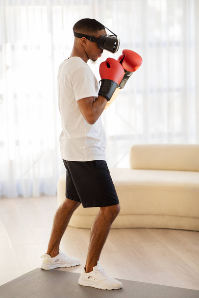 Afro-Amerikaanse man sportman in VR bril en rode bokshandschoenen met virtuele training thuis, vechter met behulp van augmented reality om te trainen voor mama of kickboxing competitie, verticale full length shot - Foto, afbeelding