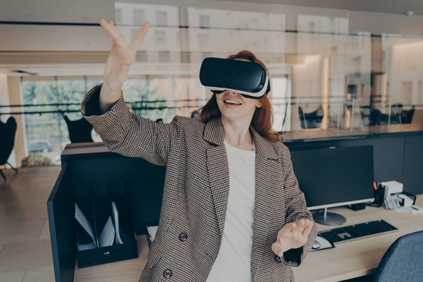 Female office worker using virtual reality to visualize and browse project files, using her hands to control objects in digital world, excited business woman wearing vr headset gesturing in office - Фото, изображение