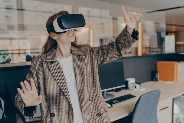 Szczęśliwa pracownica biurowa korzystająca z gry wirtualnej rzeczywistości podczas przerwy, bawiąca się przenośnymi okularami VR, poruszającymi się rękami przedmiotami w cyfrowym świecie, stojąca przed swoim miejscem pracy w słuchawkach - Zdjęcie, obraz