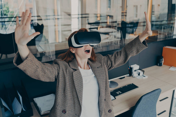 Impressionante donna impiegato con VR occhiali sulla testa vedere qualcosa di enorme e incredibile nella realtà virtuale mentre in piedi in ufficio, rendendo sorpresa espressione facciale con entrambe le mani in aria - Foto, immagini