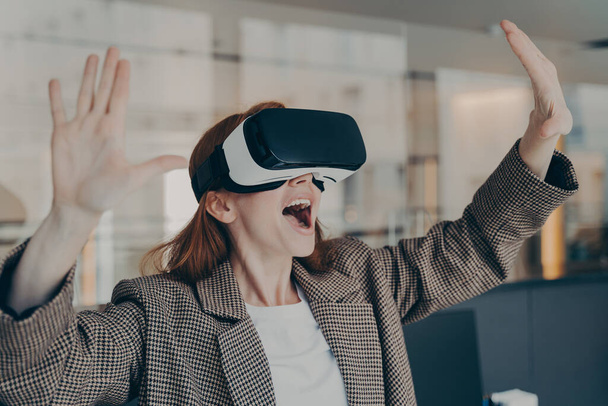 Nadšený mladý podnikatel žena zažívá virtuální realitu pomocí VR brýle, zatímco stojí v úřadu, vyjadřuje emoce s gestem a šťastný výraz obličeje, nemůže uvěřit tomu, co vidí - Fotografie, Obrázek