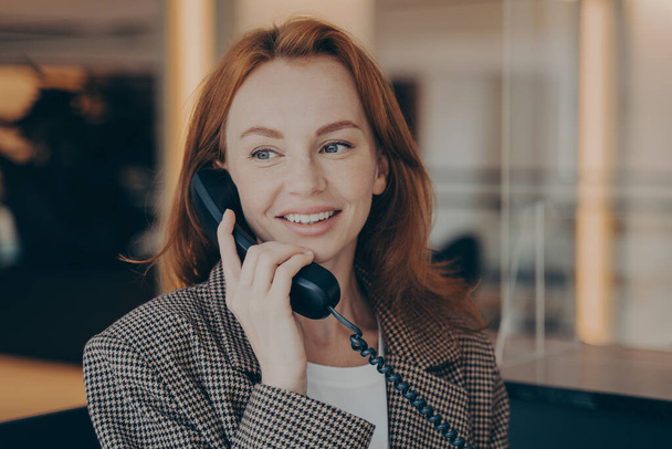 Portrait d'une employée de bureau satisfaite aux cheveux roux appelant un partenaire d'affaires ou un client au travail, utilisant un téléphone fixe noir et un environnement de bureau souriant et flou en arrière-plan - Photo, image