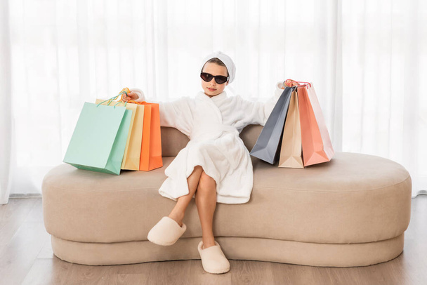 Cool Little Girl Wearing Bathrobe And Stylish Sunglasses Holding Bright Shopping Bags While Sitting On Couch At Home, Shopaholic Female Child Enjoying Fashion And Seasonal Sales, Copy Space - Foto, afbeelding