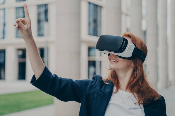 VR технології для вашого бізнесу. Щаслива молода бізнес-леді, використовуючи окуляри віртуальної реальності, стоїть на відкритому повітрі, керує проектом пальцем, тестує гарнітуру, офісні будівлі на розмитому фоні
 - Фото, зображення