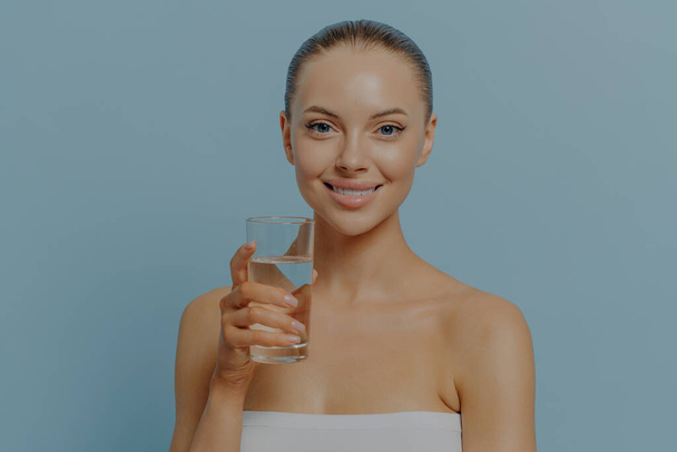 Водний баланс. Молода щаслива жінка з чистою шкірою тримає склянку чистої води і посміхається на камеру, позуючи в студії з голими плечима, ізольовані на синьому. Догляд за тілом, харчування та охорона здоров'я
 - Фото, зображення
