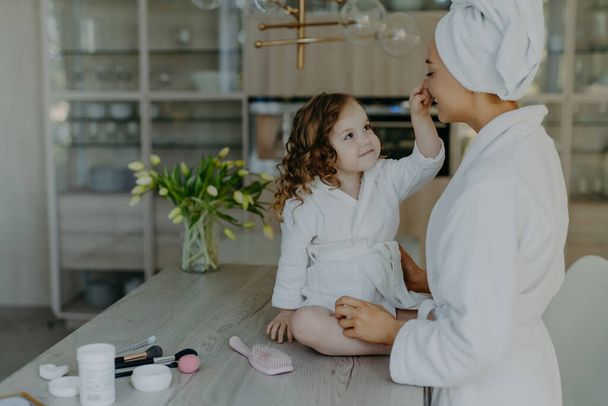 Vrij krullend klein meisje raakt neus van haar moeder gekleed in witte zachte dressing toga zit op tafel met cosmetische producten gaan make-up te doen voor moeder samen tegen gezellige interieur - Foto, afbeelding