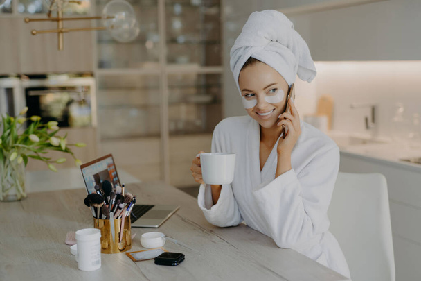 Happy young European woman undergoes beauty procedures after morning shower wears hydrogel moisturising patches dressed in white bathrobe drinks aromatic coffee talks via mobile phone poses indoor - Foto, imagen