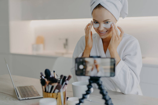 Photo of woman blogger influencer records video how to apply beauty pads under eyes looks at smartphone camera teaches followers doing skin care procedure poses against home interior in bathrobe - Foto, afbeelding