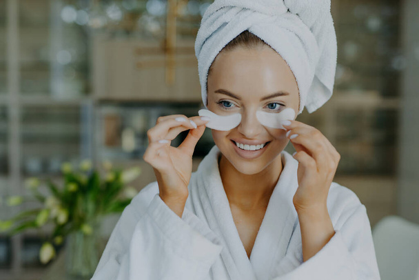 Close up shot of happy lovely European woman takes off beauty patches smiles gently has refreshed well groomed complexion wears bathrobe and wrapped towel undergoes cosmetic procedures at home - Foto, Imagem