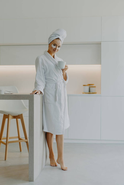 Full length shot of refreshed young European woman poses bare feet in bathrobe at kitchen drinks coffee in morning undergoes beauty procedures before going to work enjoys domestic atmosphere - 写真・画像