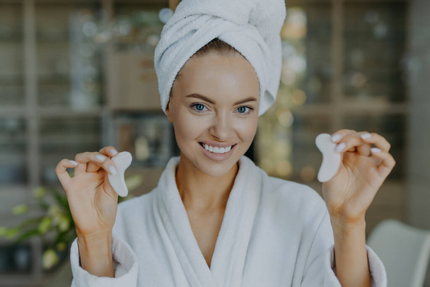 Boldog európai nő tartja hidrogél foltok a kezében mosolyog foggal egészséges friss bőr pozitívan néz ki a kamera használ kozmetikai termék, hogy távolítsa el a sötét karikák a szem alatt visel fürdőköpenyt. - Fotó, kép