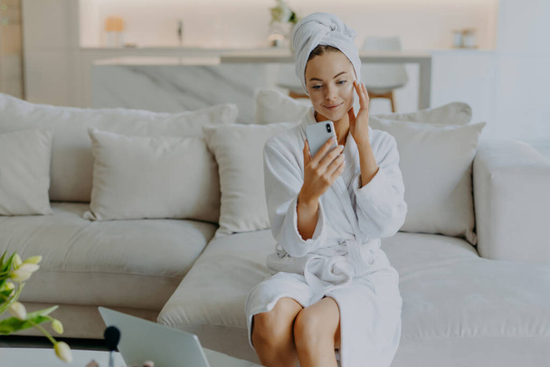 Gorgeous lady touches face gently applies face cream makes video call holds smartphone in front of herself wears bathrobe and towel sits on comfortable sofa against cozy home interior. Beauty concept - Photo, Image