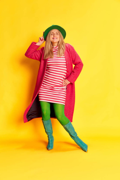 Happiness, smiling. Portrait of woman in colorful clothes, hat, coat and green tights posing against yellow studio background. Concept of emotions, facial expression, lifestyle and fashion - Fotó, kép