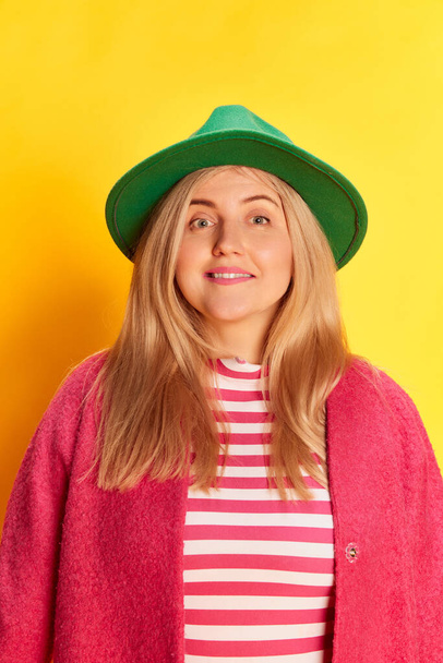 Humor positivo. Retrato de mujer en ropa colorida, sombrero verde y abrigo rosa posando sobre fondo amarillo estudio. Concepto de emociones, expresión facial, estilo de vida y moda - Foto, Imagen
