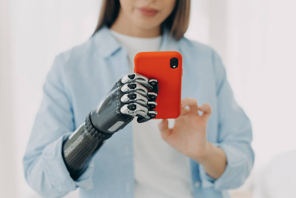 Hands of young european woman with smartphone. Girl is holding the phone with bionic artificial arm and texting. Attractive caucasian woman with artificial limb at home. Modern bionic prosthesis. - Photo, Image