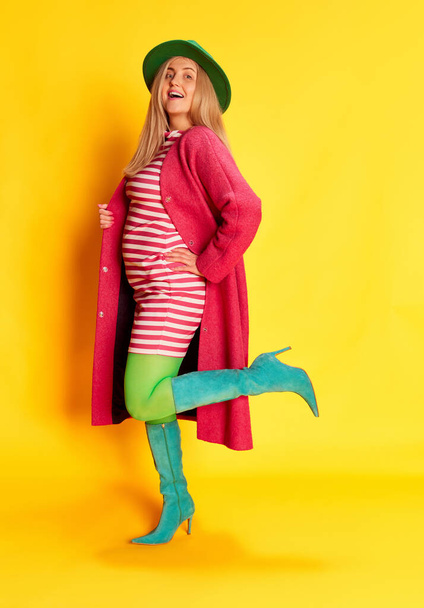 Positive mood. Portrait of woman in colorful clothes, green hat, pink coat and heeled boots posing against yellow studio background. Concept of emotions, facial expression, lifestyle and fashion - Foto, imagen