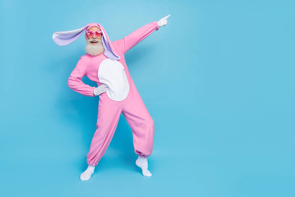 Portrait of good mood elderly pensioner wear pink bunny costume indicating empty space hand on waist isolated on blue color background. - Фото, изображение