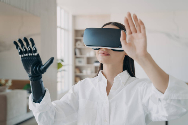 Happy handicapped girl in vr glasses at home. Disabled person gets rehabilitation. Young european woman with artificial hand is excited with 3d cyber vision. Amputee has modern bionic limb. - Foto, afbeelding