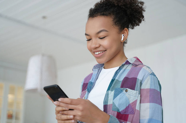 Music listening in airpods on mobile phone. Joyful teenage african american girl enjoying favourite song in wireless earphones. Student is browsing internet and social media on smartphone. - Foto, Imagen