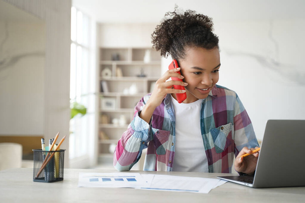 Modern biracial teen girl student discuss learning task with classmate by phone call looking at laptop. Teenager schoolgirl talking with friend while doing homework sitting at desk at home. - Photo, Image