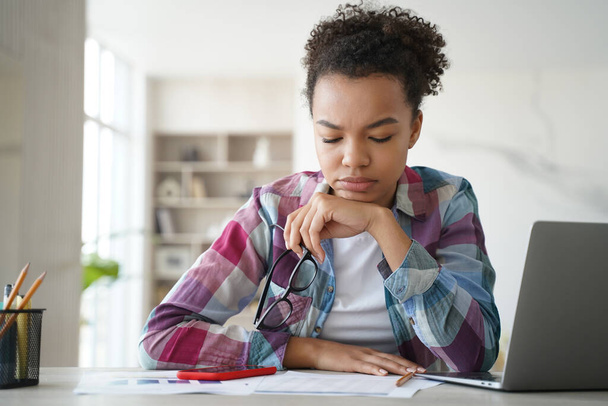 Focused biracial schoolgirl studying at desk with laptop, preparing for exam. Pensive tired mixed race teen girl student doing homework, learning task at home. Distance education concept. - 写真・画像