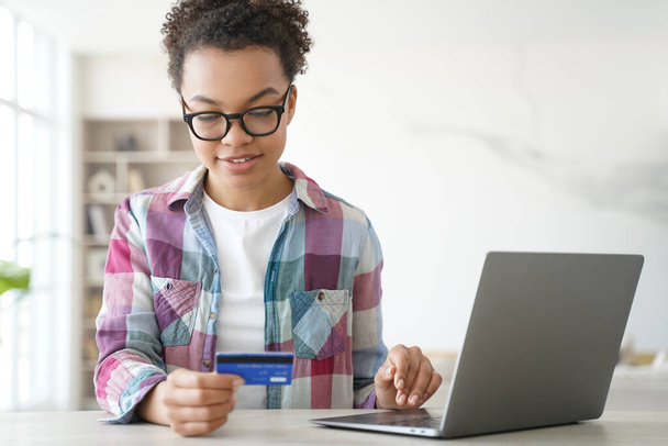 Африканская американская девочка-подросток держит кредитную карту покупок онлайн на ноутбуке. Молодая двуличная женщина покупает оплату в Интернете на компьютере дома, просматривает банковские услуги, проверяет бонус наличными. - Фото, изображение