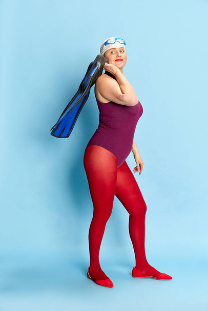 Full-length image of woman in swimming suit and red tights posing with flippers against blue studio background. Time for sport. Concept of emotions, facial expression, lifestyle and fashion - Foto, Bild