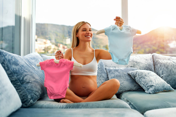 A beautiful young pregnant woman holding pink and blue baby clothes, guessing who the boy or girl will born, is sitting on the couch at home. The concept of motherhood and pregnancy. - Foto, Bild