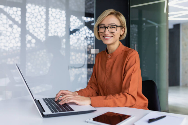 Portrait of happy and successful female programmer inside office at workplace, worker smiling and looking at camera with laptop blonde businesswoman is satisfied with results of achievements at work - Zdjęcie, obraz
