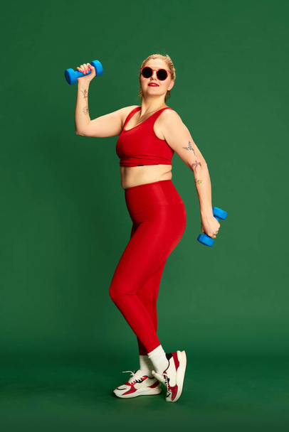 Training with gym equipment. Full-length portrait of woman in red sportive clothes posing with dumbbells against green studio background. Concept of emotions, facial expression, sport and weight loss - Foto, Bild