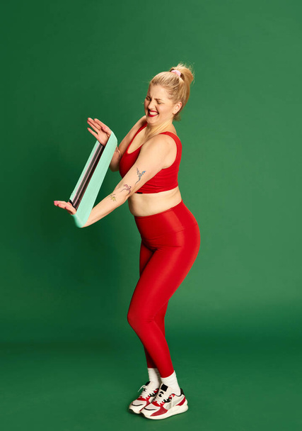 Full-length portrait of woman in red sportive clothes posing, training with fitness elastic bands against green studio background. Concept of emotions, facial expression, sport and weight loss - Foto, Bild