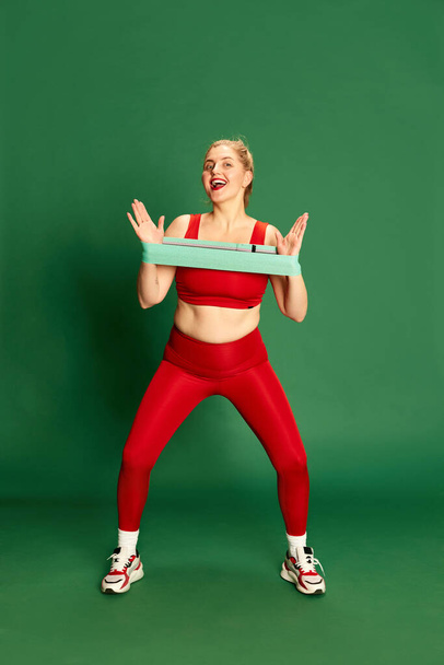 Full-length portrait of woman in red sportive clothes posing, training with fitness elastic bands against green studio background. Concept of emotions, facial expression, sport and weight loss - Φωτογραφία, εικόνα