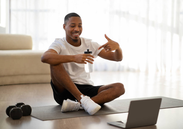 スポーツウェアフィットネスコーチのスポーツウェアの何千年もの黒人男性は、家庭でヨガマットに座って、ノートパソコンの画面と笑顔を見て、水でボトルを指して、オンラインレッスンを持っている、スペースをコピー - 写真・画像